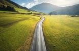 A winding road runs through green fields toward distant mountains under a bright, partly cloudy sky.